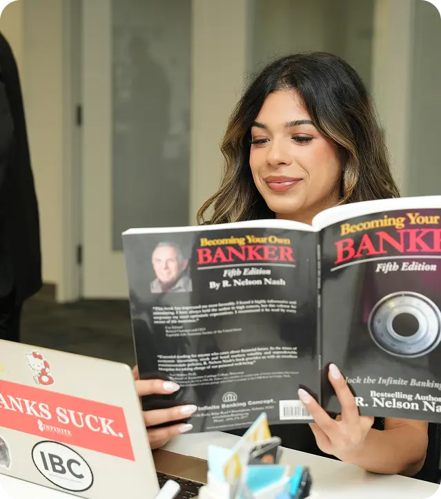 Woman with long hair reading the book 'Becoming Your Own Banker' at a desk with a laptop.