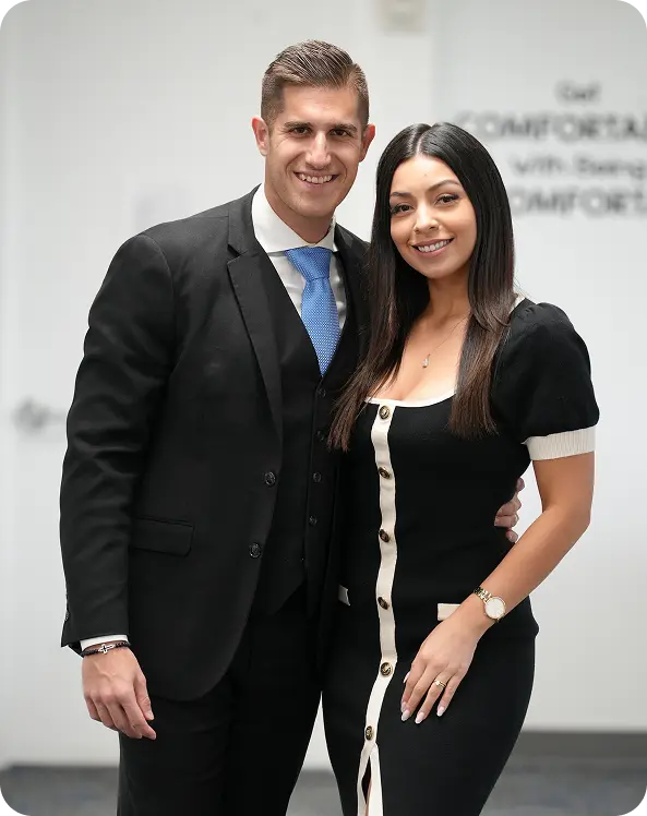 Smiling man in a suit with blue tie and woman in black dress with white buttons posing indoors.