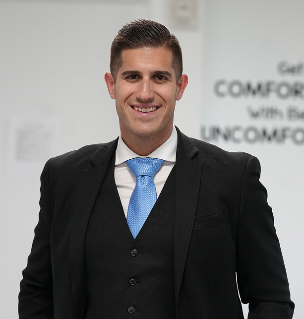 Smiling man in a black suit, white shirt, and blue tie standing indoors with a blurred motivational quote behind him.