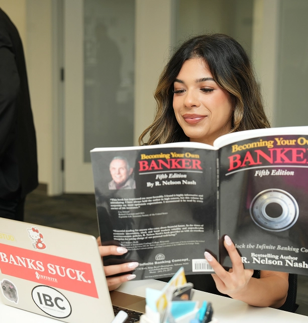 Woman reading the book 'Becoming Your Own Banker' by R. Nelson Nash at a desk with a laptop.