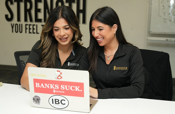 Two women in black Infinite Financial shirts smiling and looking at a laptop with bold stickers on it.