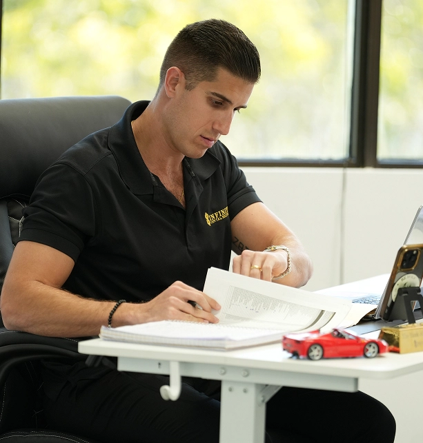Man in black polo sitting at desk, reading papers with laptop and red toy car nearby.