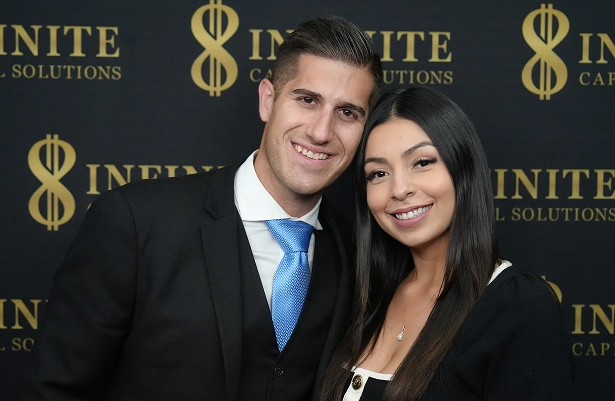 Man in a suit and woman in black blouse smiling and posing together against a branded backdrop.