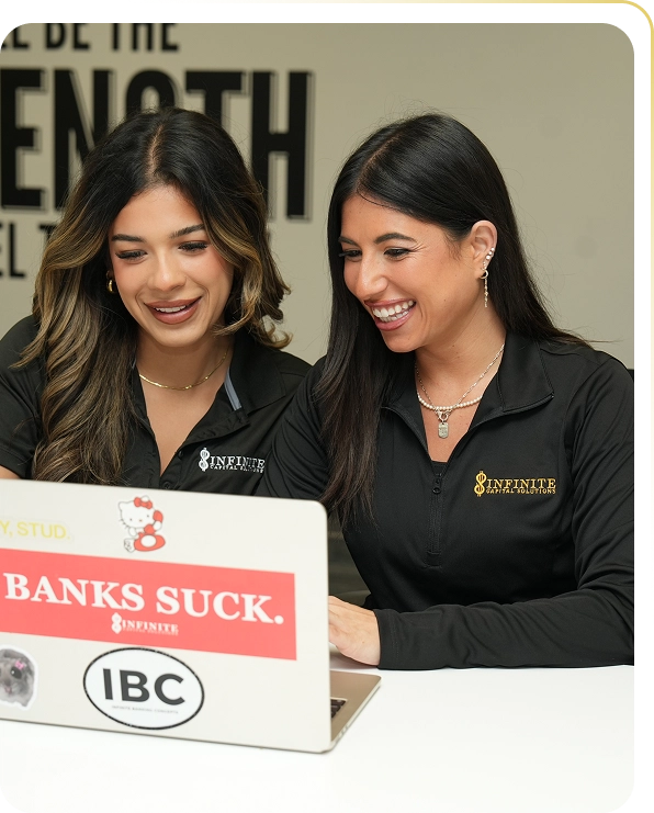 Two women wearing Infinite Capital Solutions shirts smiling and working on a laptop with stickers.