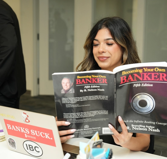 Woman reading 'Becoming Your Own Banker' book while sitting at desk with laptop and office supplies.