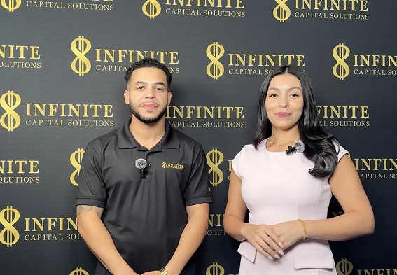 A man in a black shirt and a woman in a white dress stand before an Infinite Capital Solutions backdrop.