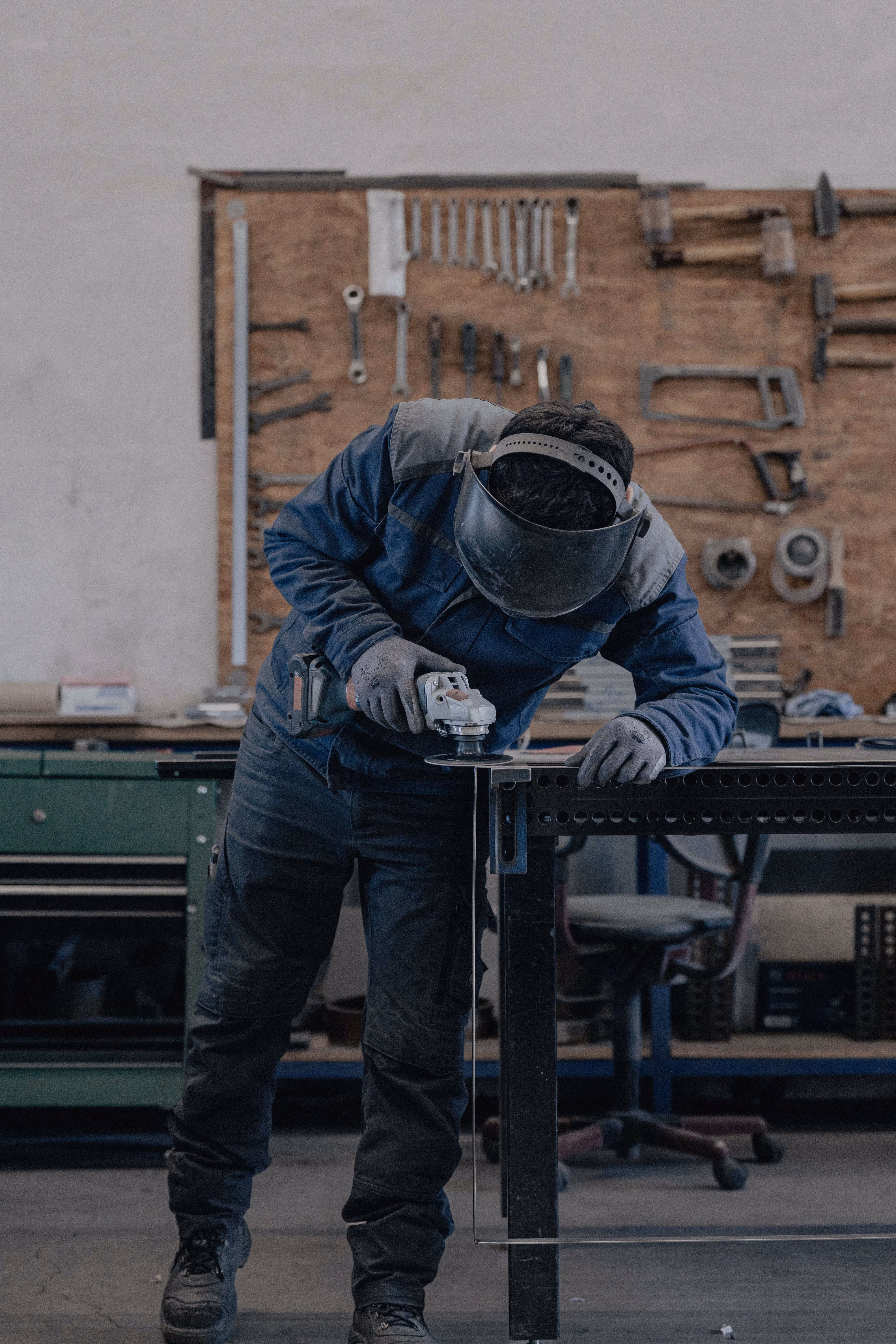 Ein Handwerker schleift Metall an einem Tisch in einer Werkstatt mit Werkzeugen an der Wand.