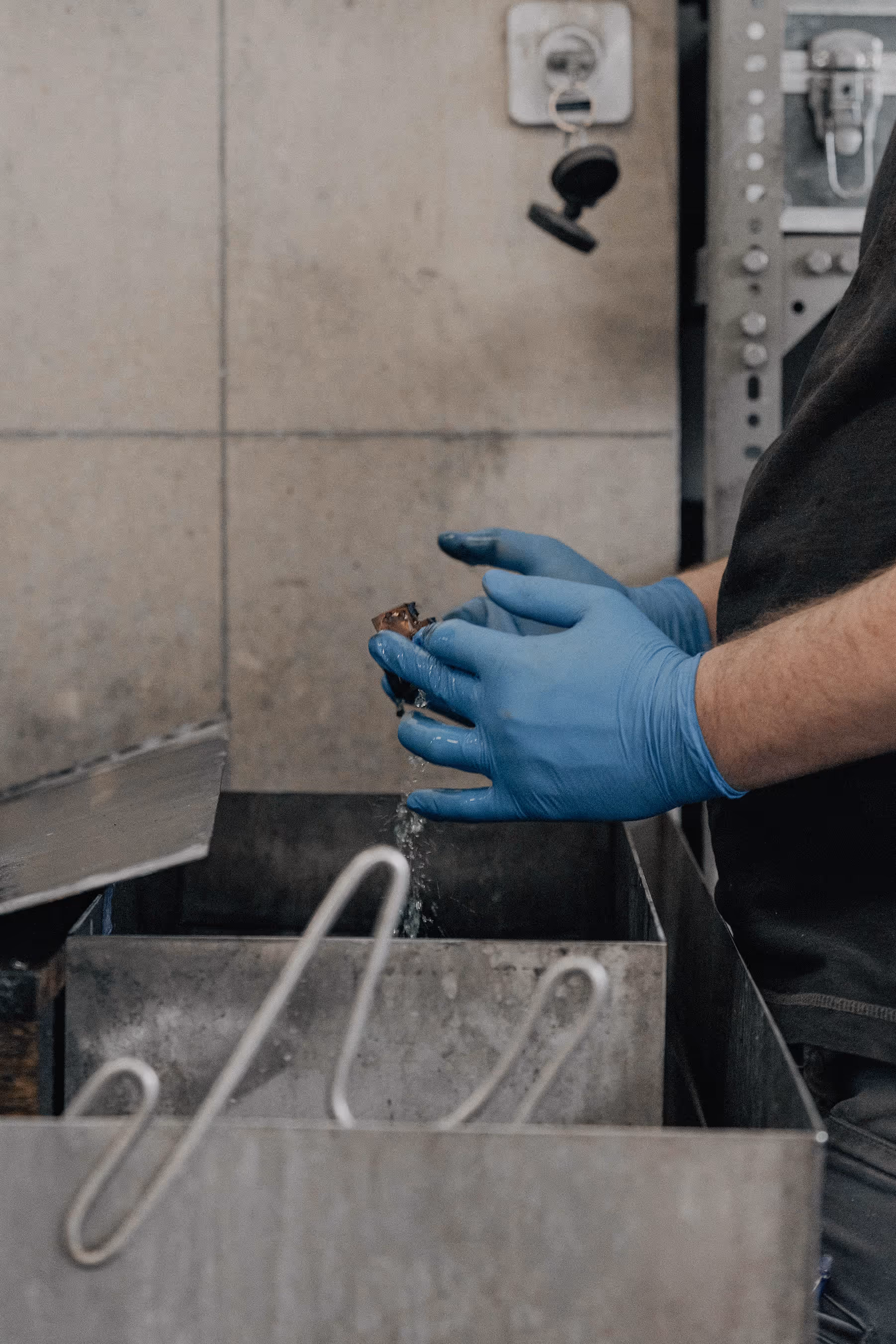 Person mit blauen Handschuhen hält ein Metallteil über eine industrielle Waschstation.