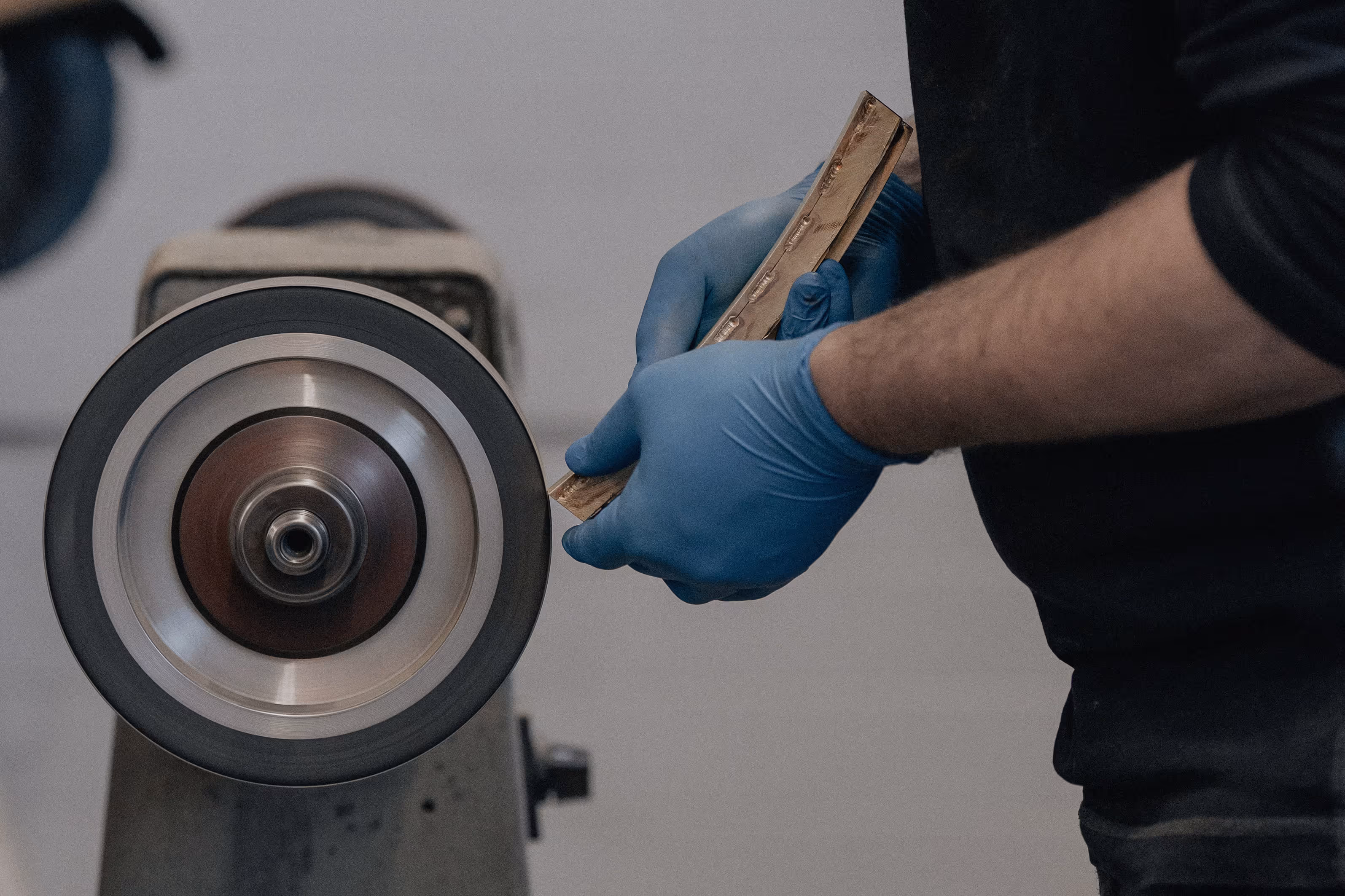 Person mit blauen Handschuhen hält Metallstück zu rotierendem Schleifrad an Maschine.
