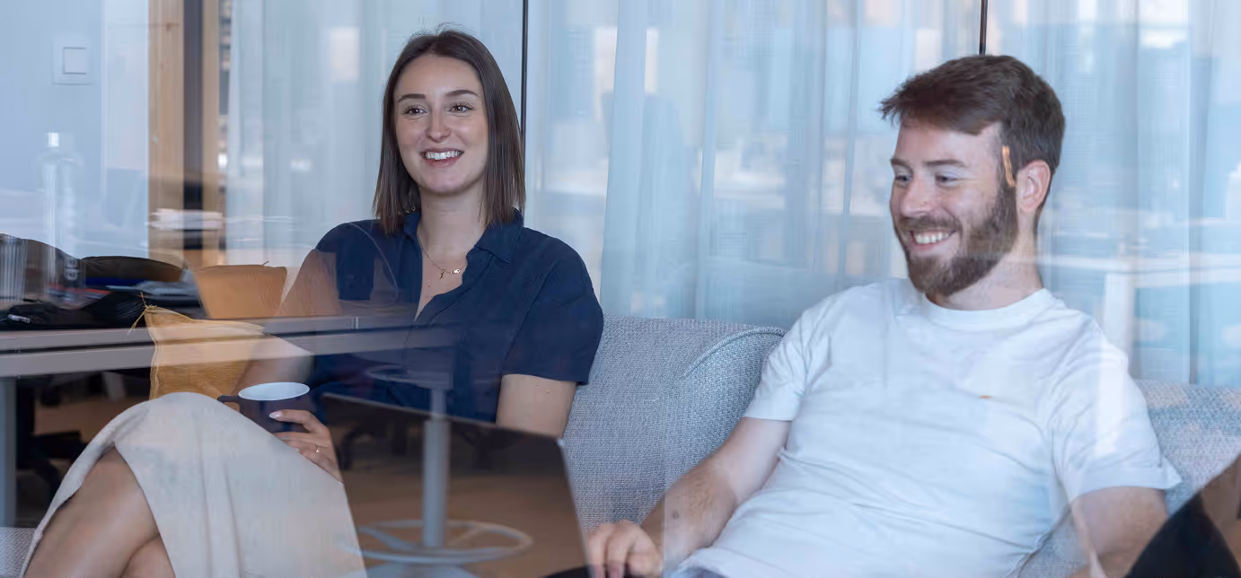 Smiling man and woman sitting on a sofa, looking at a laptop together behind a glass window.