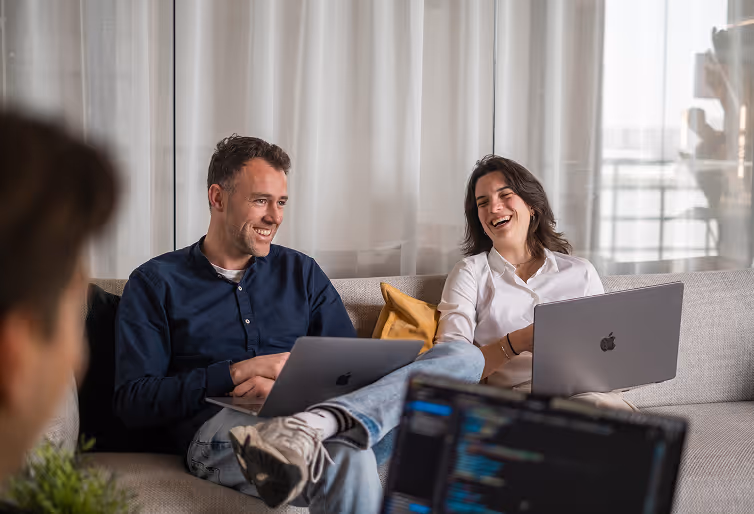 Two people sitting on a couch with laptops, smiling and chatting in a casual office setting.