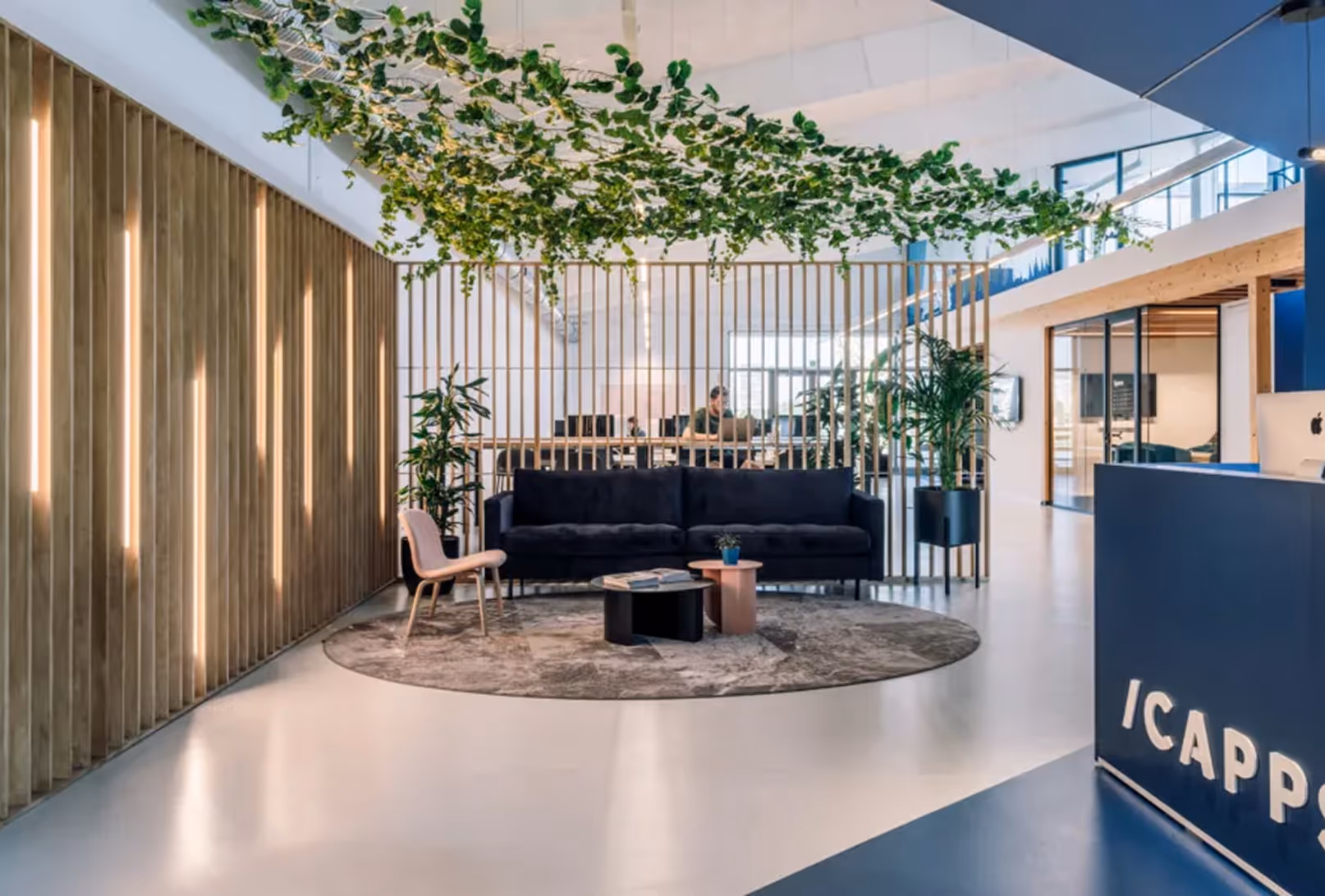 Modern office lounge with black sofa, round rug, plants, wood slats partition, and hanging greenery above.
