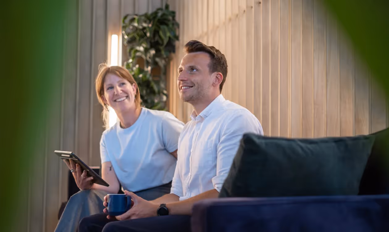 Smiling man holding a cup and woman with a tablet sitting on a couch indoors.