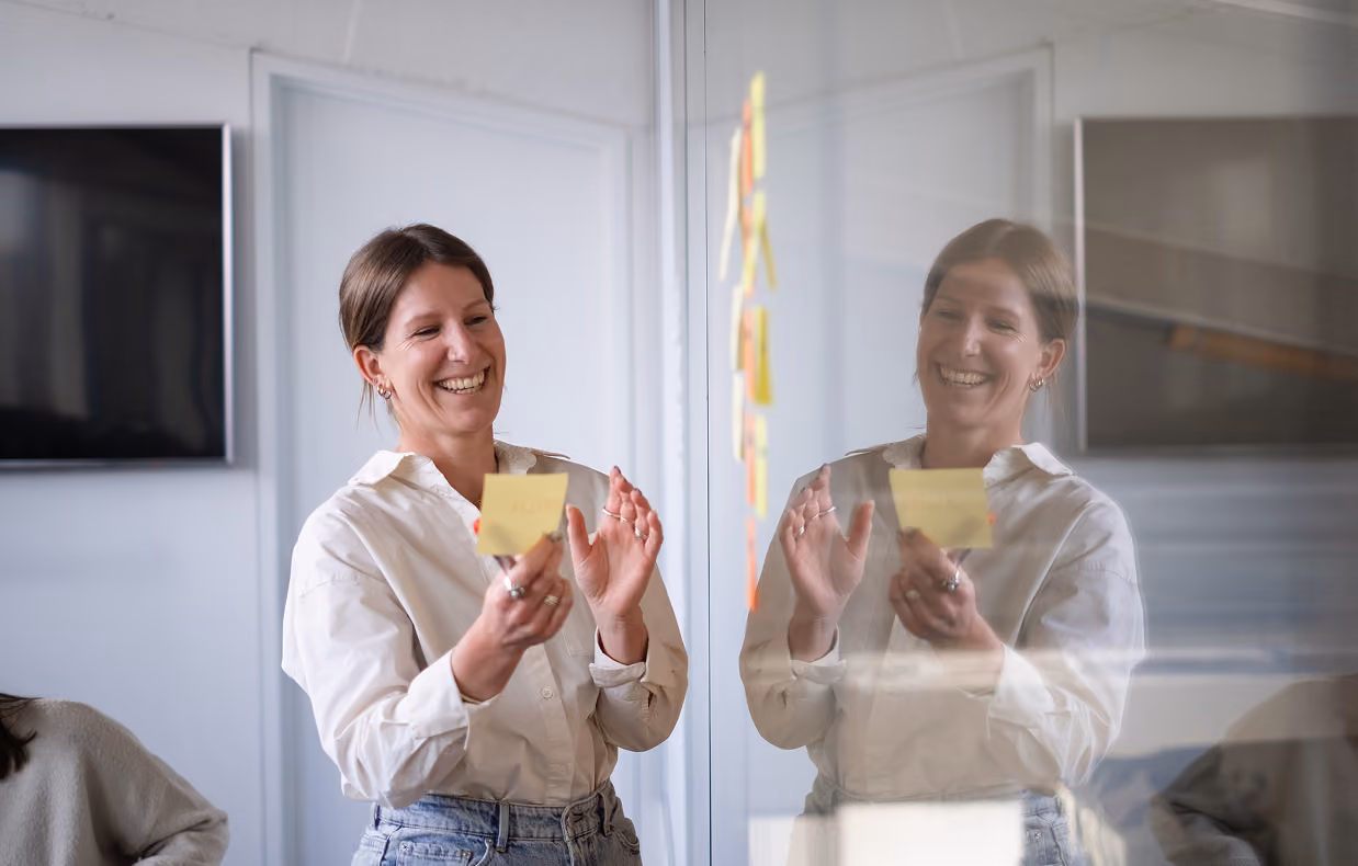 Smiling woman placing sticky notes on a glass wall during a collaborative meeting.