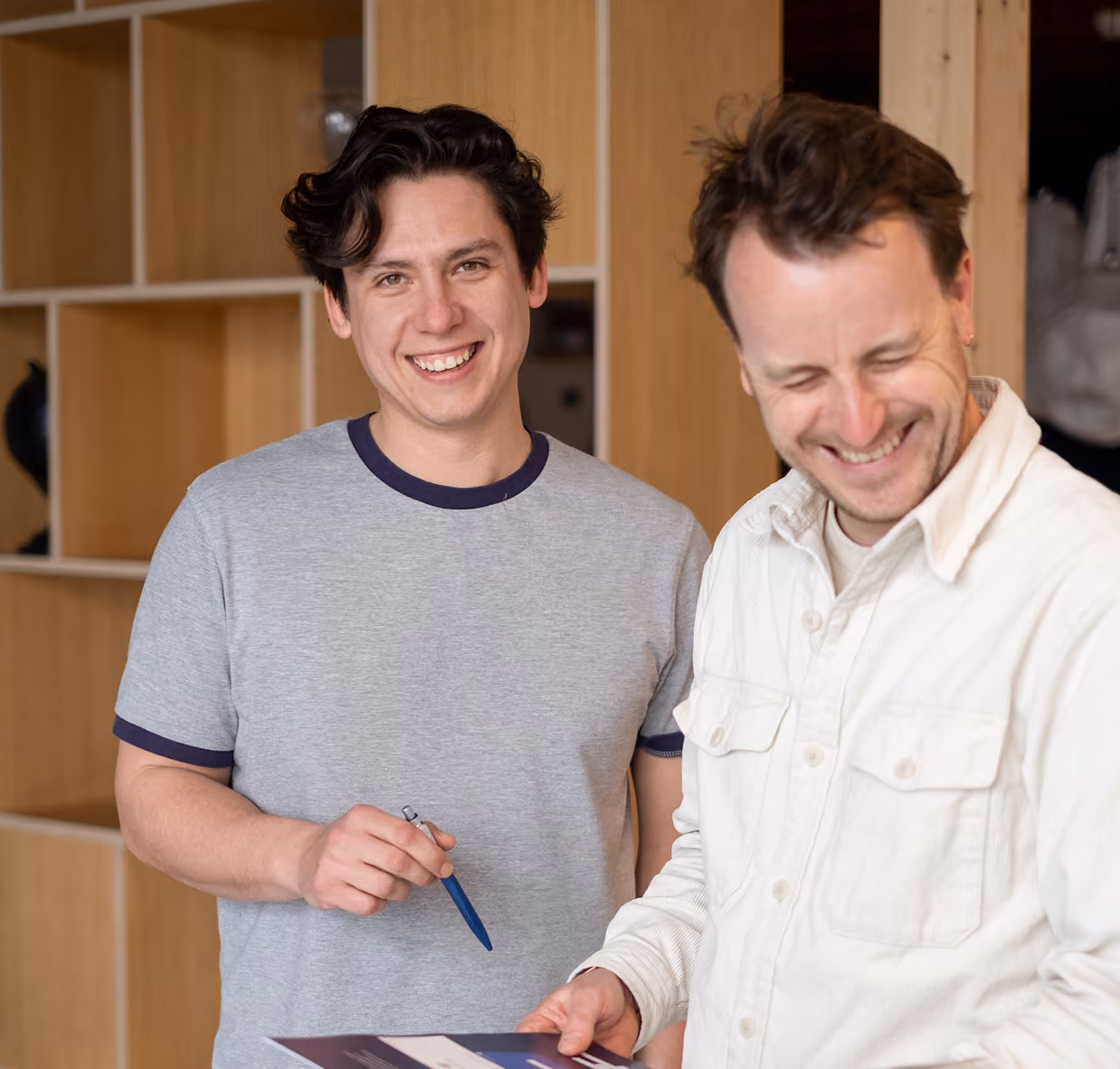 Two men smiling indoors, one holding a pen and the other holding a brochure or paper.