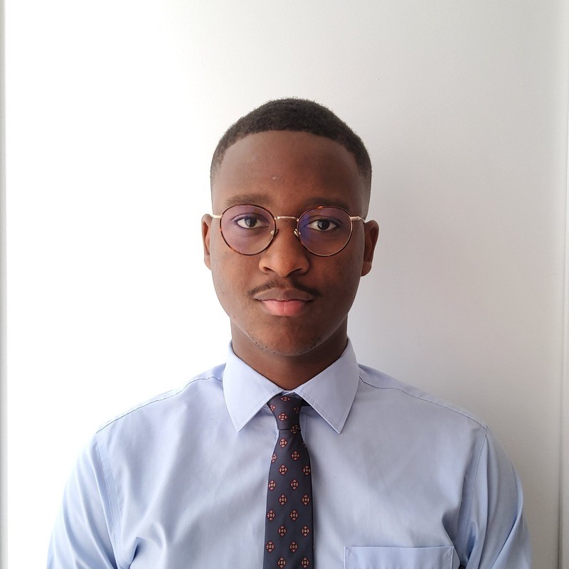 Young man with short hair and round glasses wearing a light blue shirt and patterned tie.
