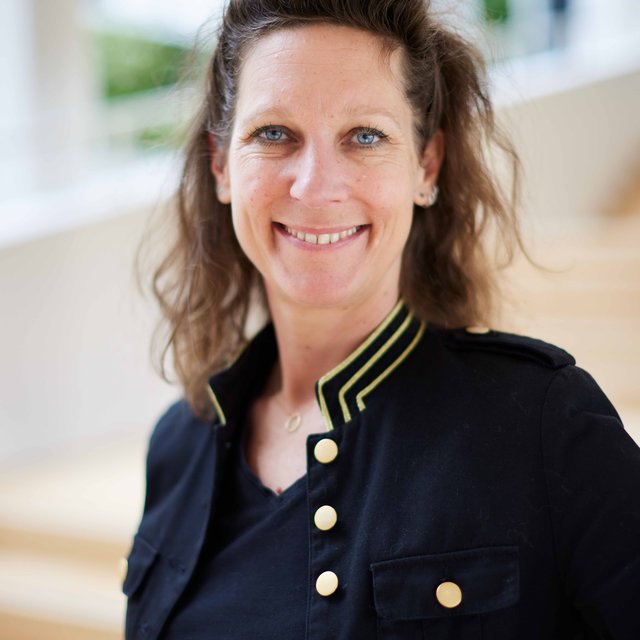 Smiling woman with curly brown hair wearing a black jacket with gold buttons and trim.