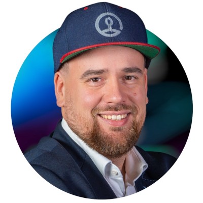 Smiling man with a beard wearing a blue cap and dark jacket on a colorful blurred background.