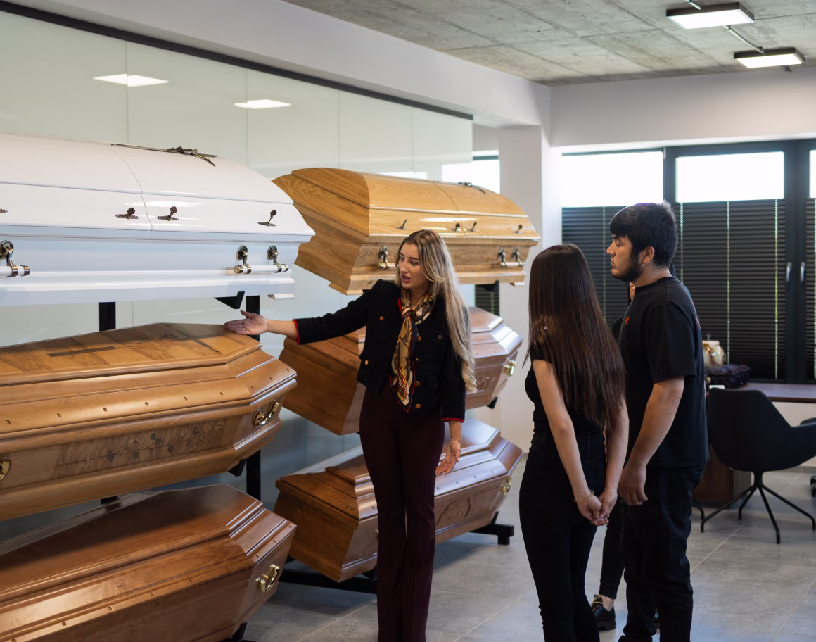 Funeral director showing wooden and white coffins to a couple in a funeral home showroom.