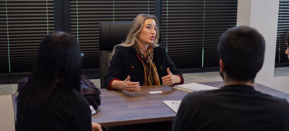 Woman speaking to two people across a table in a meeting room with blinds closed.