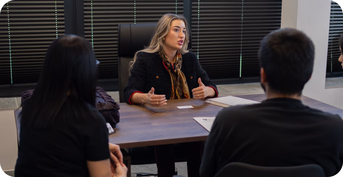 Businesswoman in a black jacket speaking to a group around a conference table in an office.