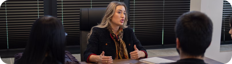 Woman speaking and gesturing during a meeting with two others in an office with blinds.
