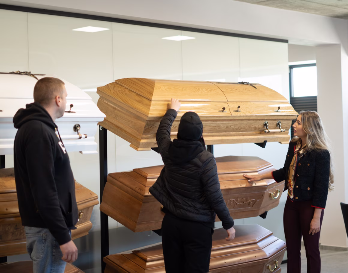 Woman showing wooden and white coffins to a couple in a showroom with large windows.