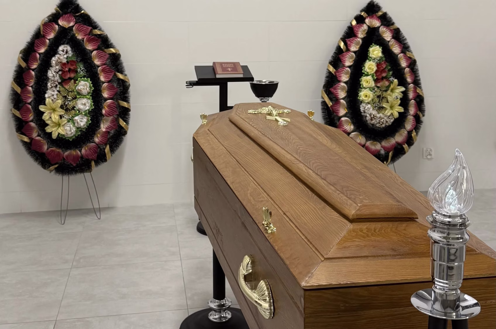Wooden coffin with cross, flanked by transparent chairs and floral wreaths in a minimal room.