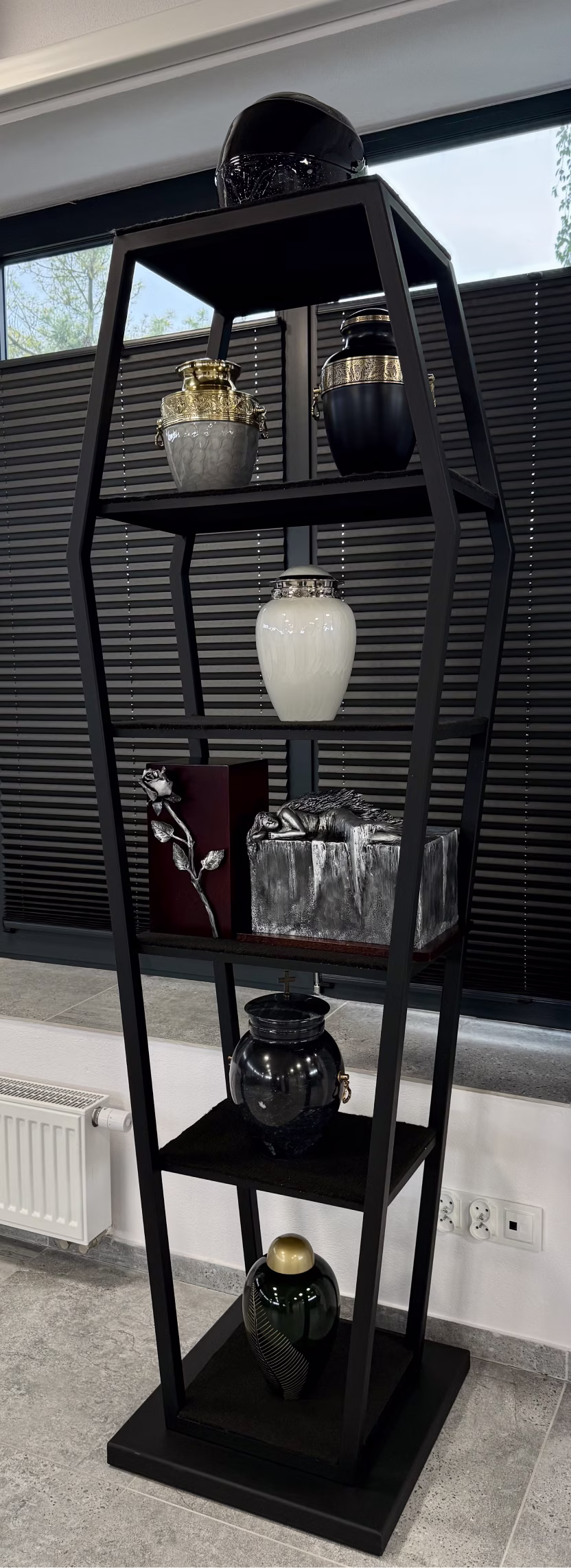 Black metal coffin-shaped shelving unit holds urns, a helmet, and funeral memorabilia indoors.