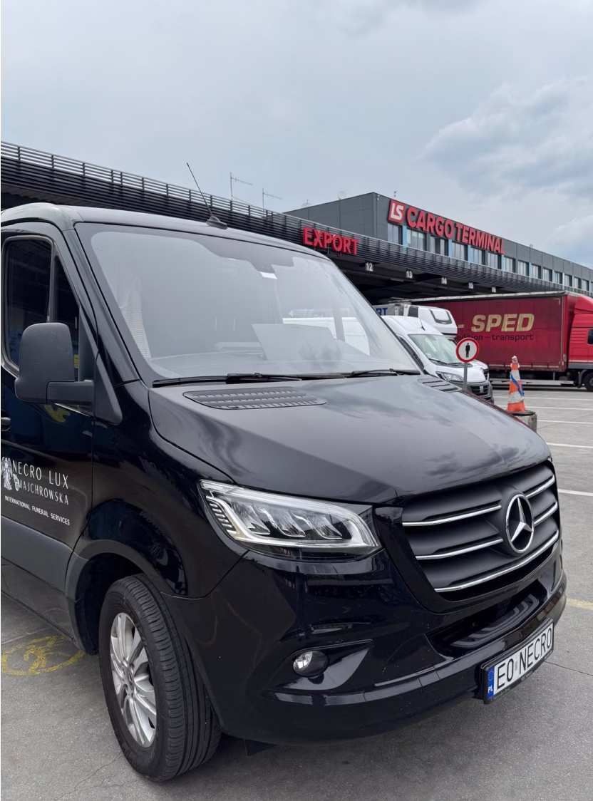 White Mercedes van with 'Necro Lux Majchrowska International Funeral Services' parked by a brick building with a gold dome.