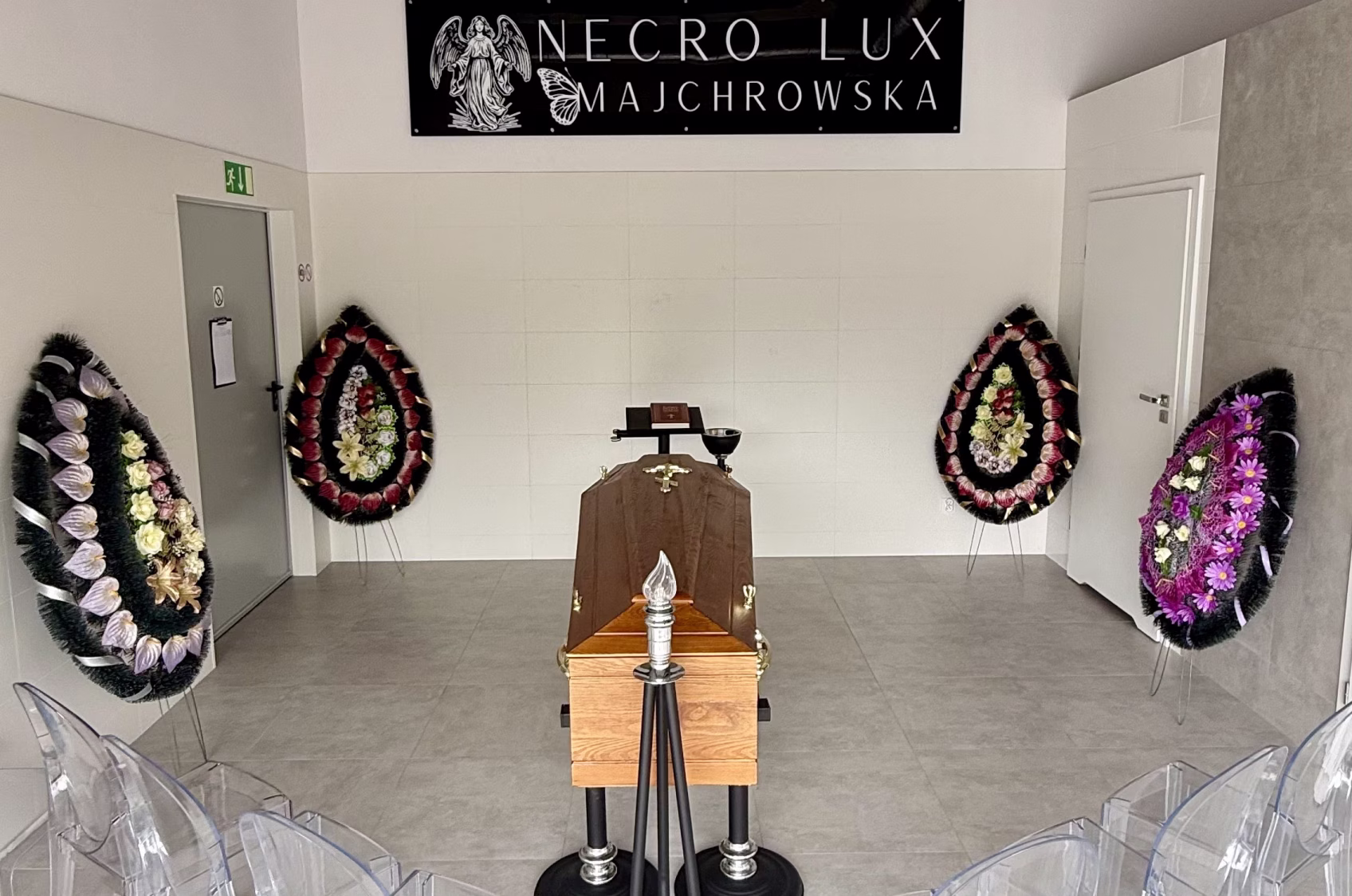 Funeral setting with wooden coffin, floral wreath, clear chairs, and a sign with an angel and butterfly.
