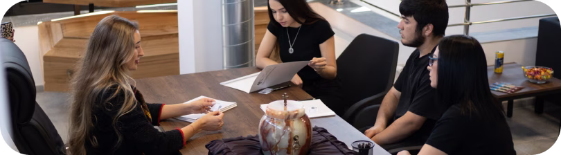 Woman speaking and gesturing during a meeting with two others in an office with blinds.