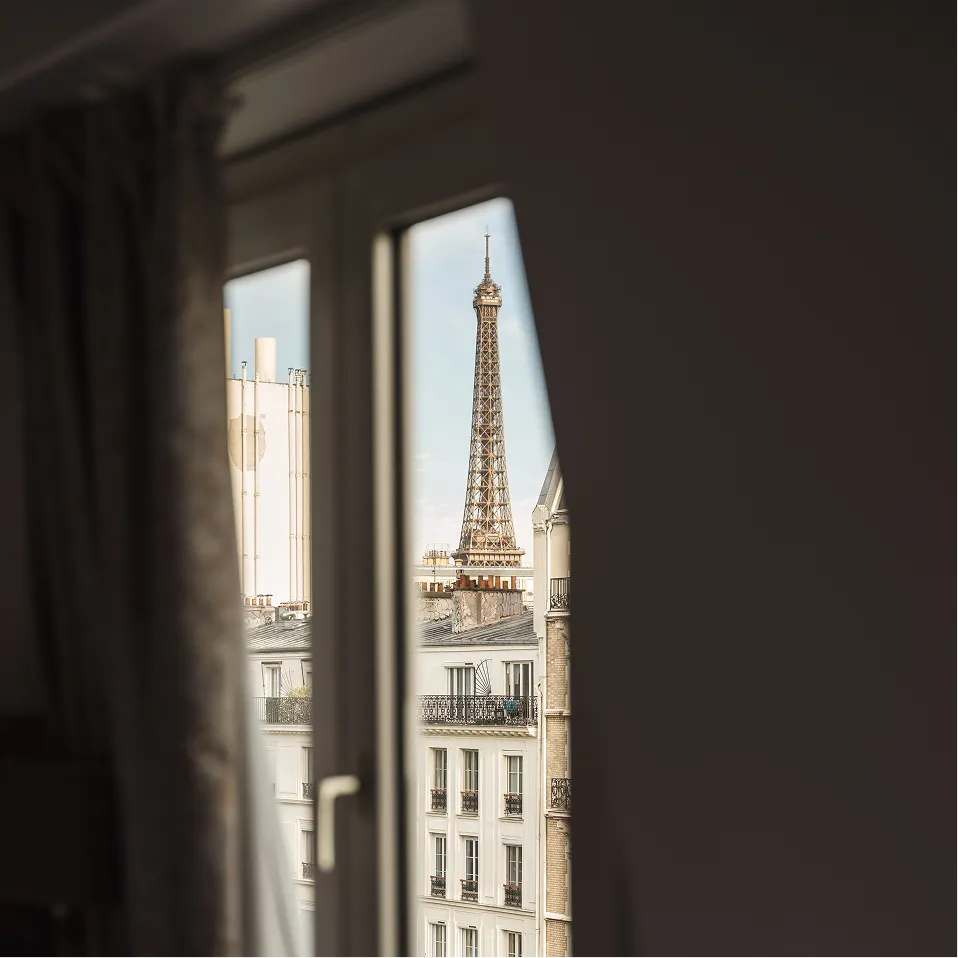 Vue de la Tour Eiffel à travers une fenêtre d'un appartement parisien avec des bâtiments blancs.
