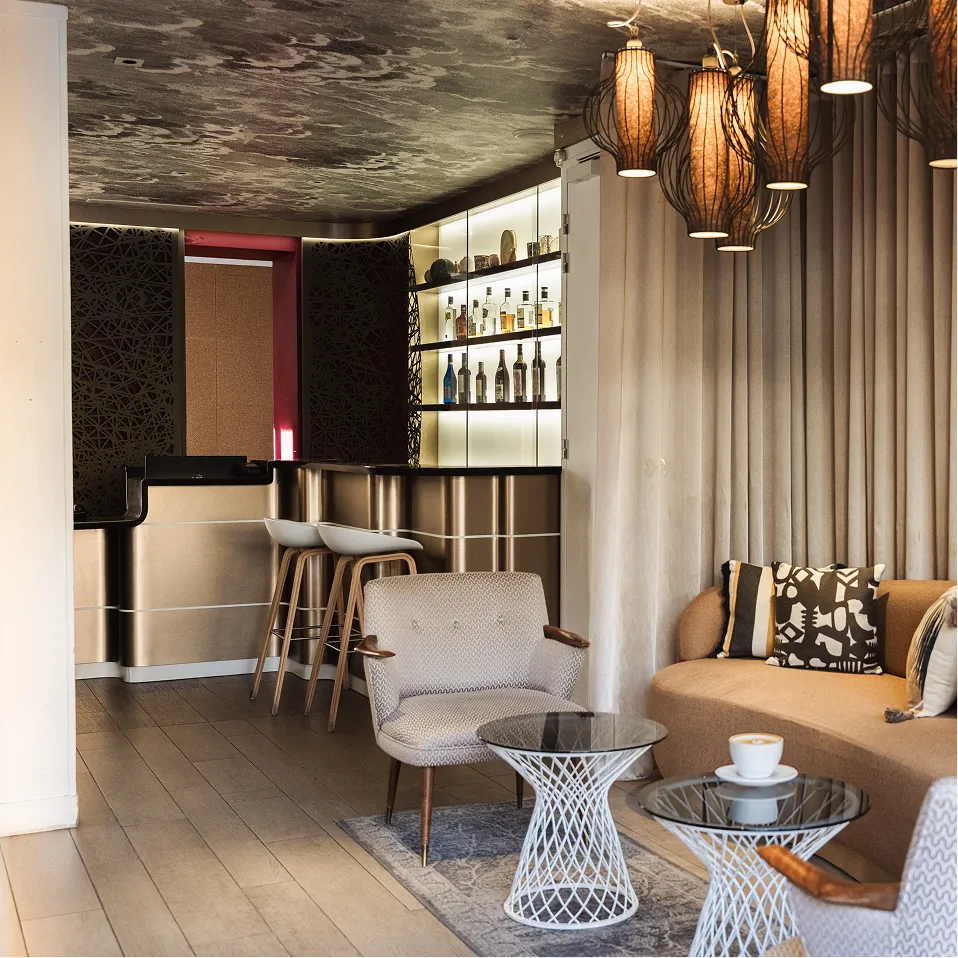 Salon avec fauteuils, canapé beige, tables basses en verre et bar éclairé avec bouteilles.
