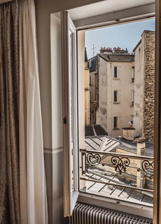 Vue depuis une fenêtre ouverte sur un balcon avec rampe en fer forgé et bâtiments denses en arrière-plan.