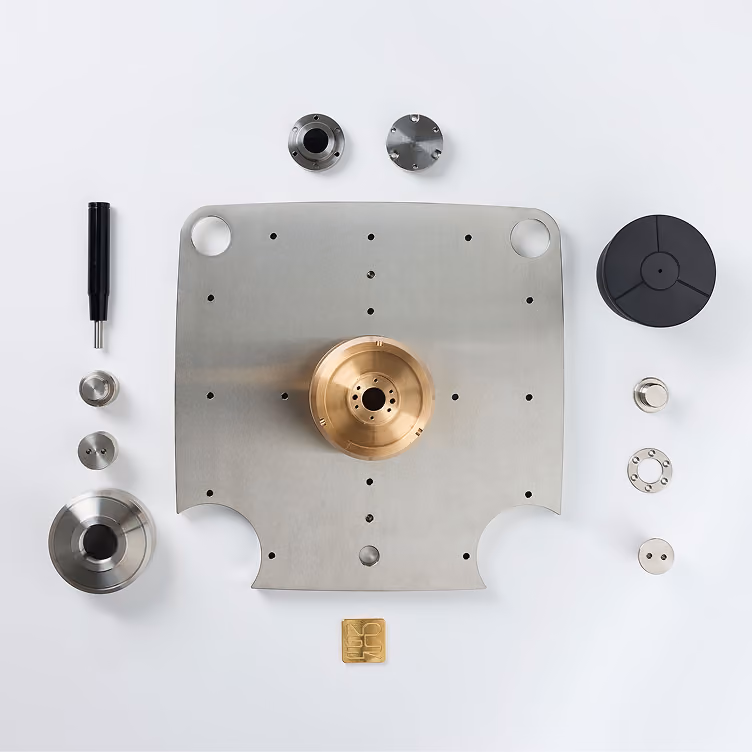 Componenti metallici di precisione disposti ordinatamente su sfondo bianco, tra cui una piastra centrale in metallo con foro circolare e un componente cilindrico dorato montato sopra, vari pezzi rotondi e cilindrici argentati e neri attorno.