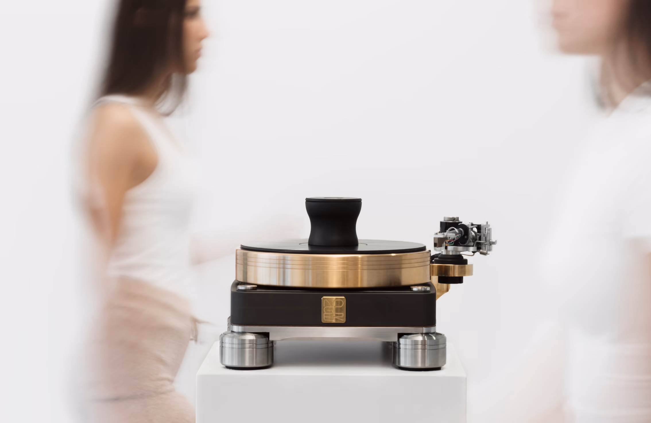 Giradischi di design moderno con struttura nera e finiture in metallo color oro, posizionato su un piedistallo bianco, con due persone sfocate che camminano ai lati.