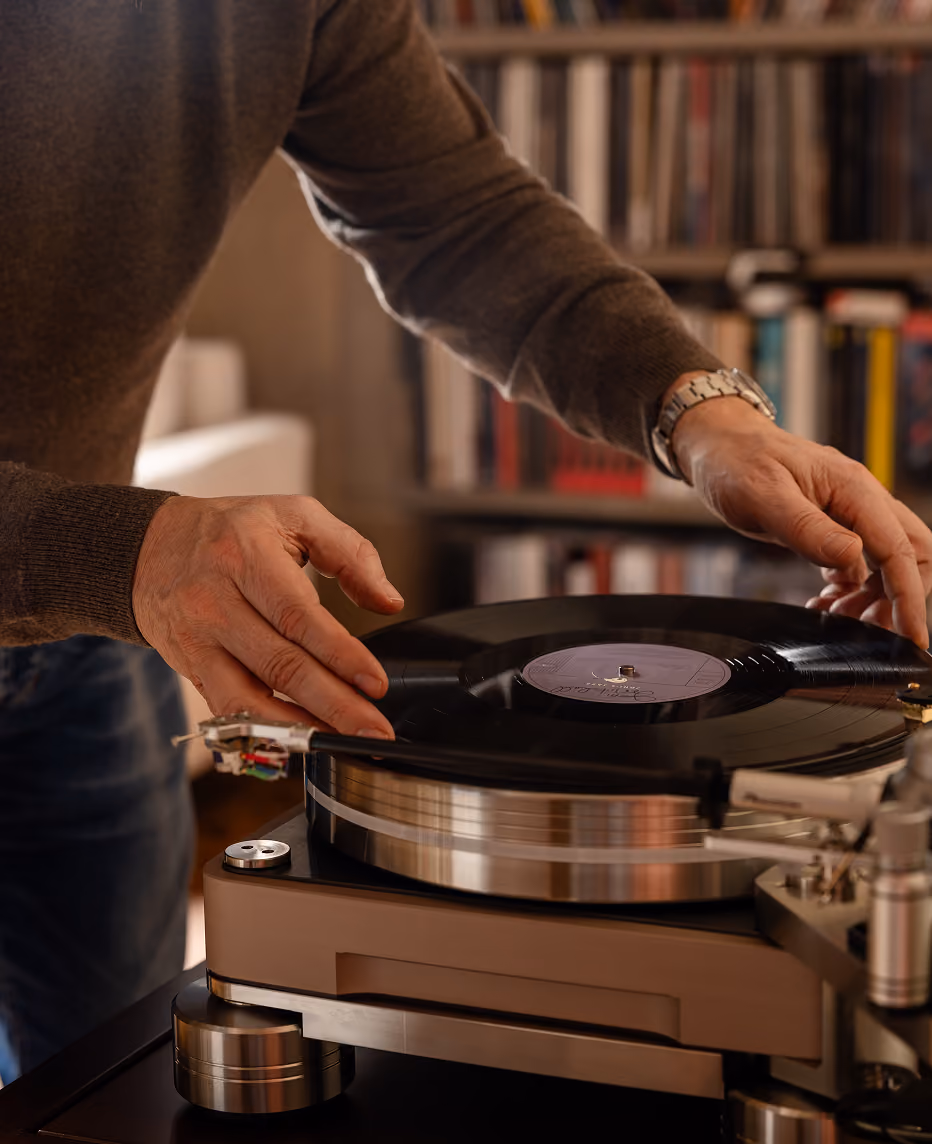 Persona che sta posizionando un disco in vinile su un giradischi in una stanza con libreria sullo sfondo.