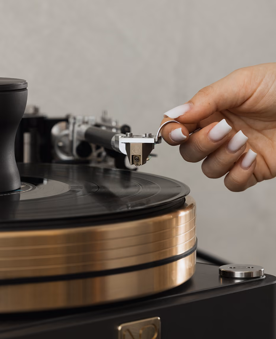 Mano con unghie curate che tiene un anello e posiziona la puntina su un giradischi con un vinile nero, sfondo sfocato grigio.