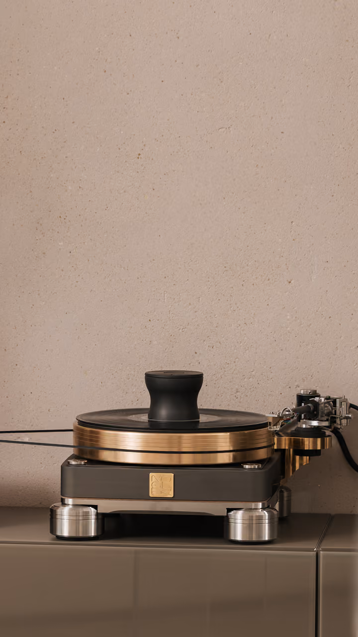 Giradischi di alta qualità con base nera, piatto in metallo dorato e pesetto centrale nero, posizionato su un mobile grigio con parete beige sullo sfondo.