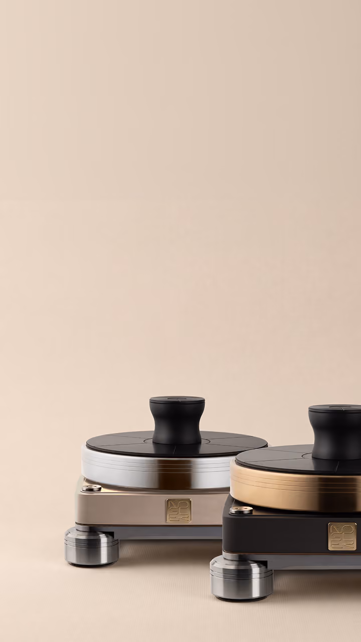 Due diffusori acustici compatti con base piatta e manopola nera sulla parte superiore, uno con dettagli in argento e l'altro con dettagli in bronzo, posizionati su una superficie beige neutra.