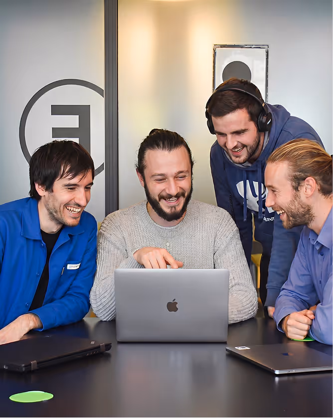 Quatre hommes souriants collaborent autour d'un ordinateur portable Apple à une table noire.