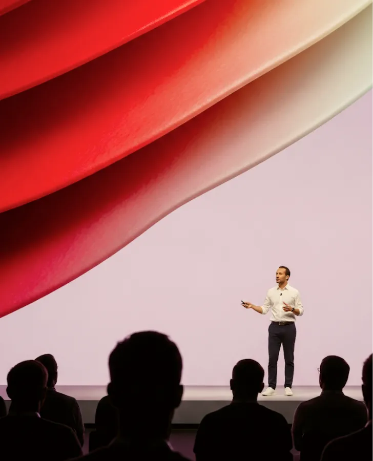Man giving a presentation on stage with abstract red and beige shapes on a large screen behind him.