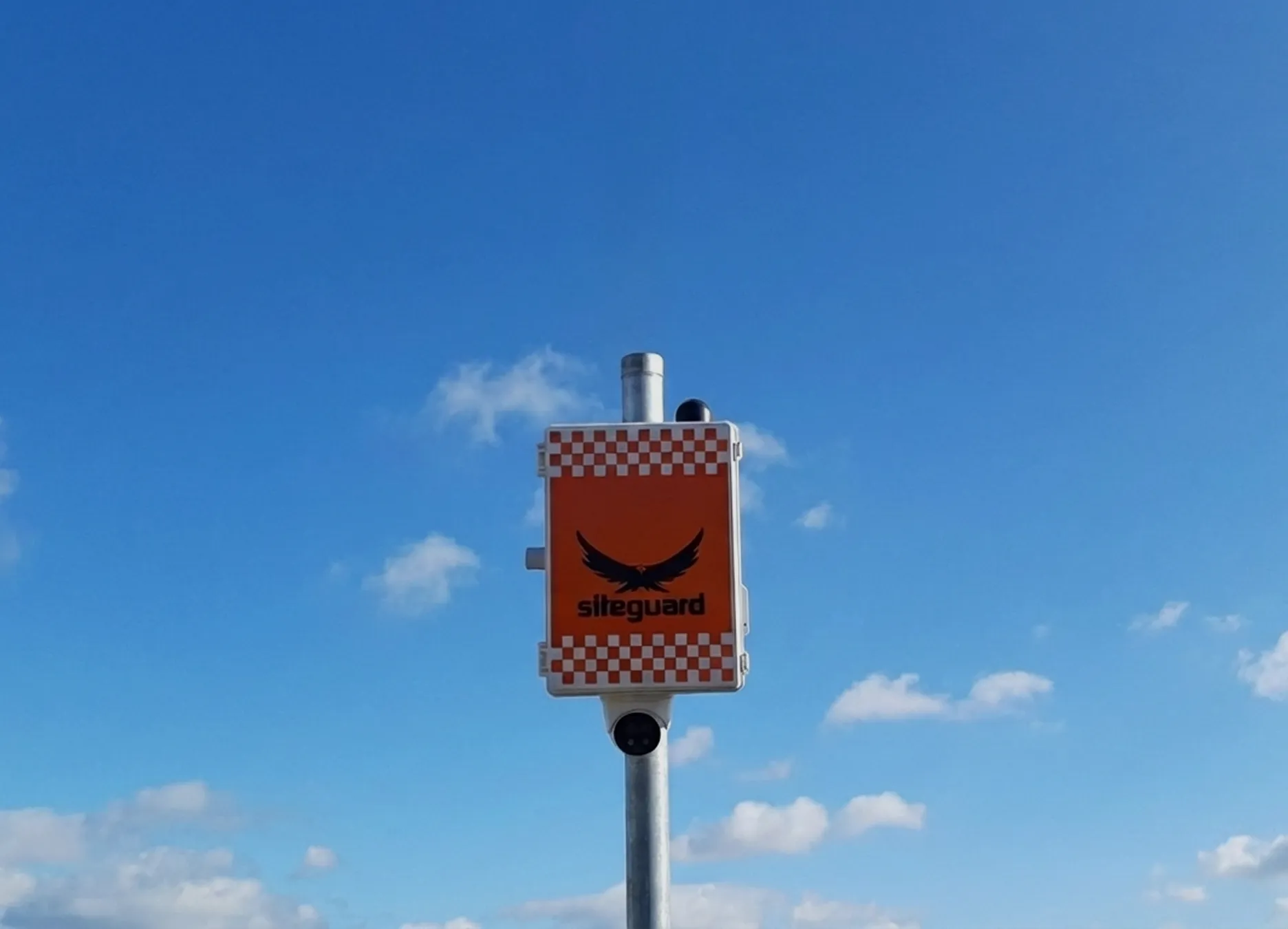 Orange siteguard safety sign with checkered border mounted on a metal pole against a blue sky.