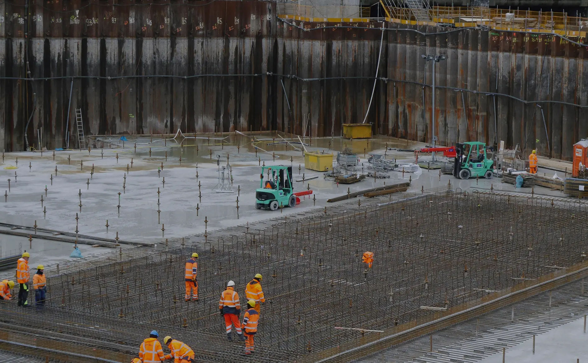 Construction workers in high-visibility clothing working on a large concrete foundation with steel rebar framework.