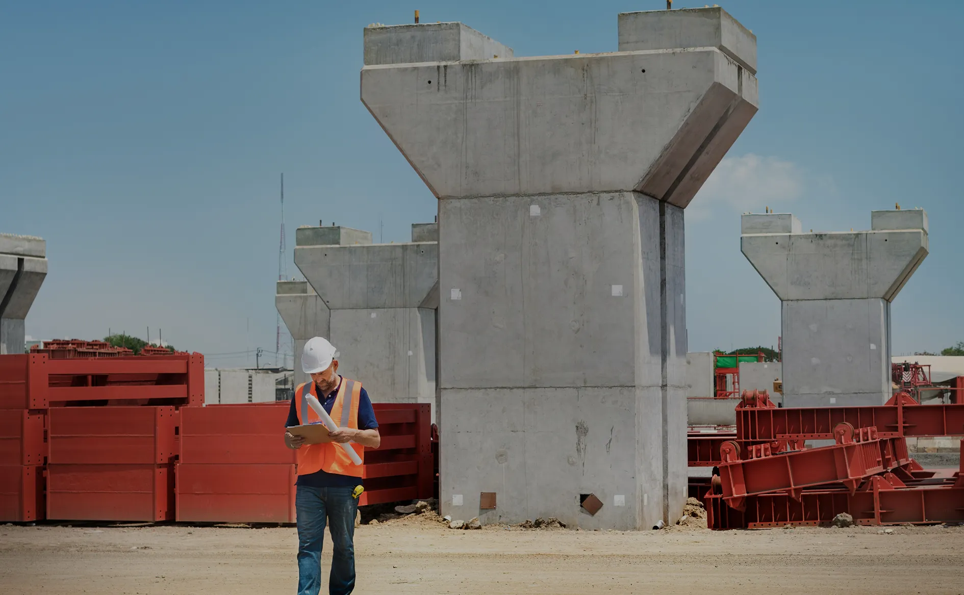 Construction worker wearing a hard hat and vest inspecting plans at a concrete bridge pillar site.