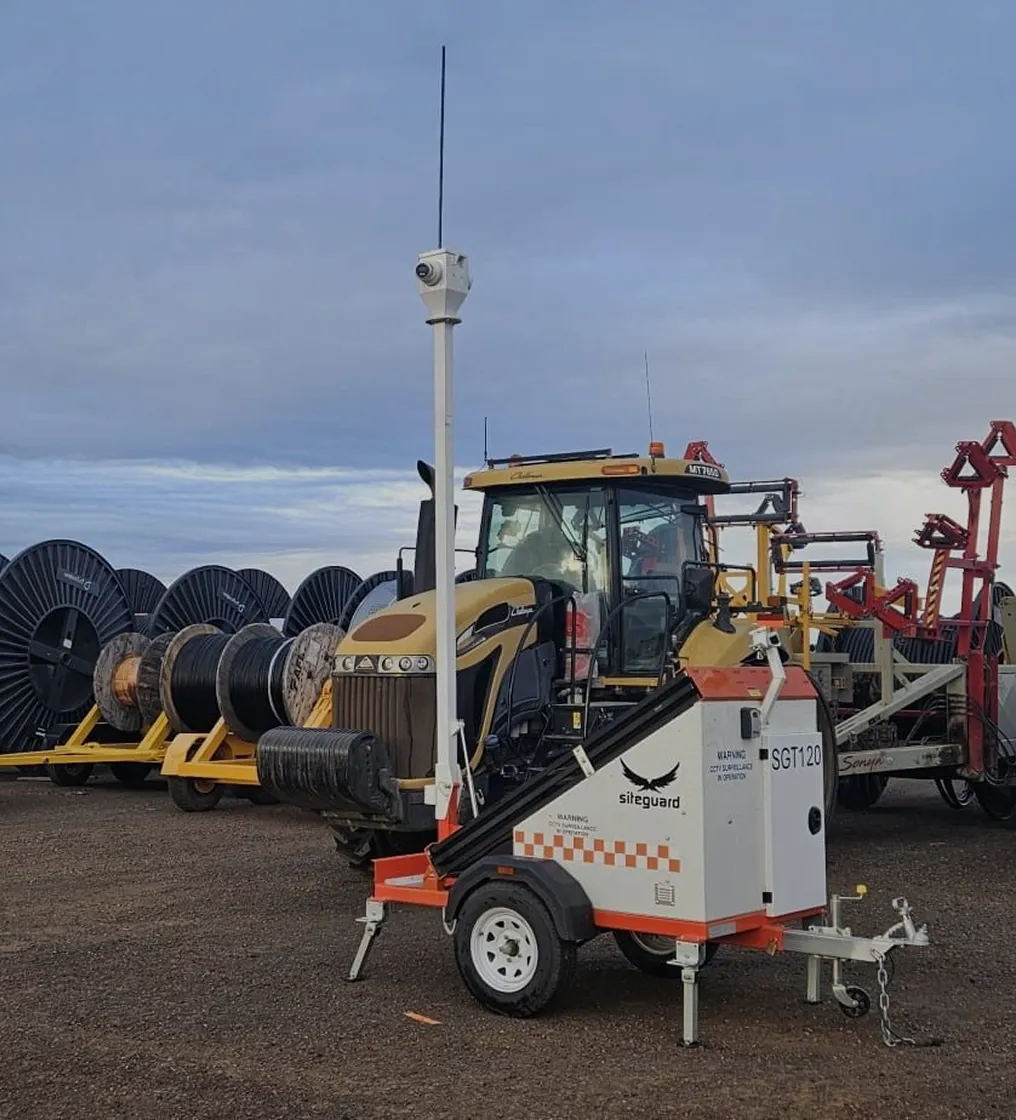Site surveillance trailer with mounted camera next to a yellow tractor and cable reels on a gravel surface.