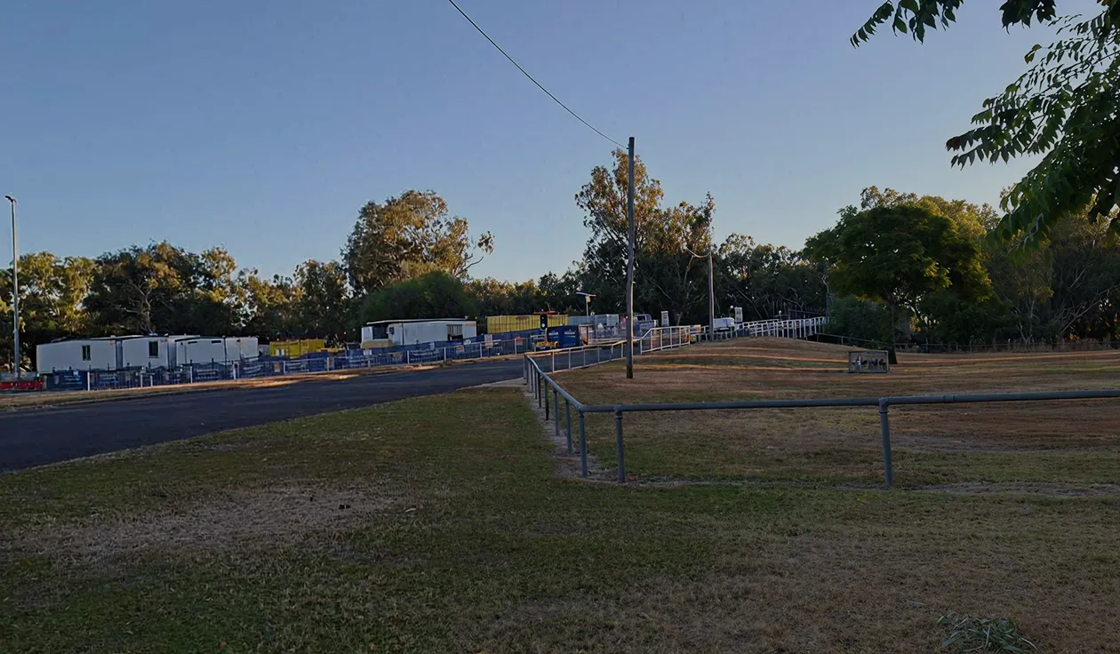 Open grassy area with scattered trees, metal fences, and a paved road under a clear blue sky.