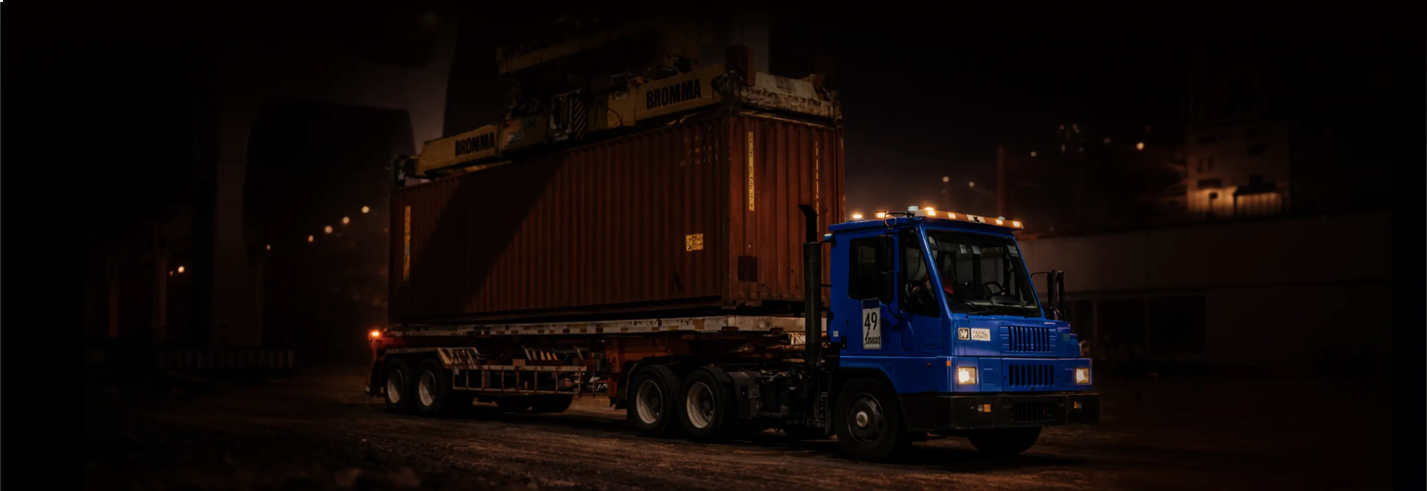 Blue truck hauling a large container on a trailer at night in an industrial area.