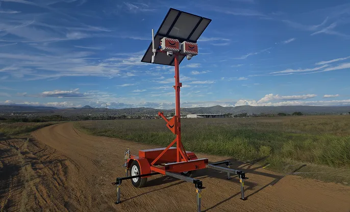 Portable solar-powered light tower mounted on a trailer parked on a dirt road in a rural area.